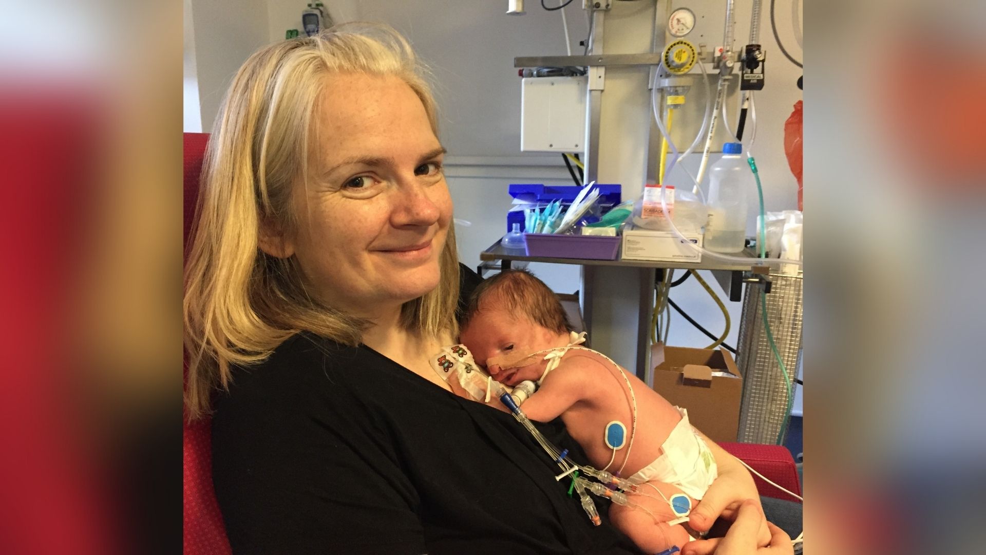 Kate, a white woman in her 30s, who has platinum blonde hair. She's wearing a black cardigan and smiling at the camera. She's holding her son William on her chest, a newborn baby who has treacher collins syndrome. There's hospital equipment in the background and William is attached to various machines.