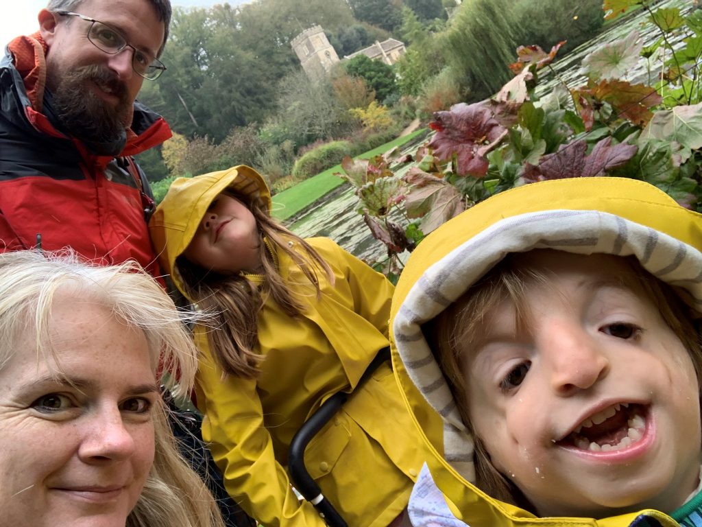 Kate, a white woman in her 30s, who has platinum blonde hair. She's looking at the camera and surrounded by her family: Nick, her husband, a white man in his 30s with brown hair, glasses and a big beard. He's wearing a red waterproof jacket. Beside him is their daughter Alex, 6-8 years old, who's wearing a yellow raincoat and leaning her head back. In the foreground is Nick and Kate's son, William, a 4-year-old boy who's smiling at the camera and has a condition called Treacher Collins Syndrome. He's wearing a yellow rain coat.