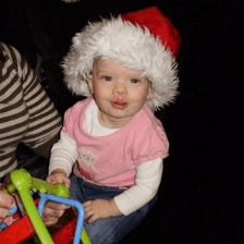 A baby wearing a Santa hat and a pink T-shirt holds a green, blue and red toy