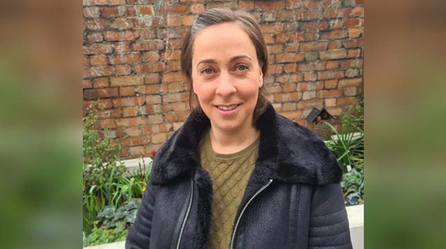 A woman with short brown hair, wearing a black jacket and green jumper, stands in front of a wall and flower bed