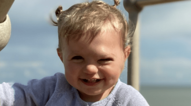 A young child wearing a grey cardigan smiles at the camera. The sky is in the background behind her.