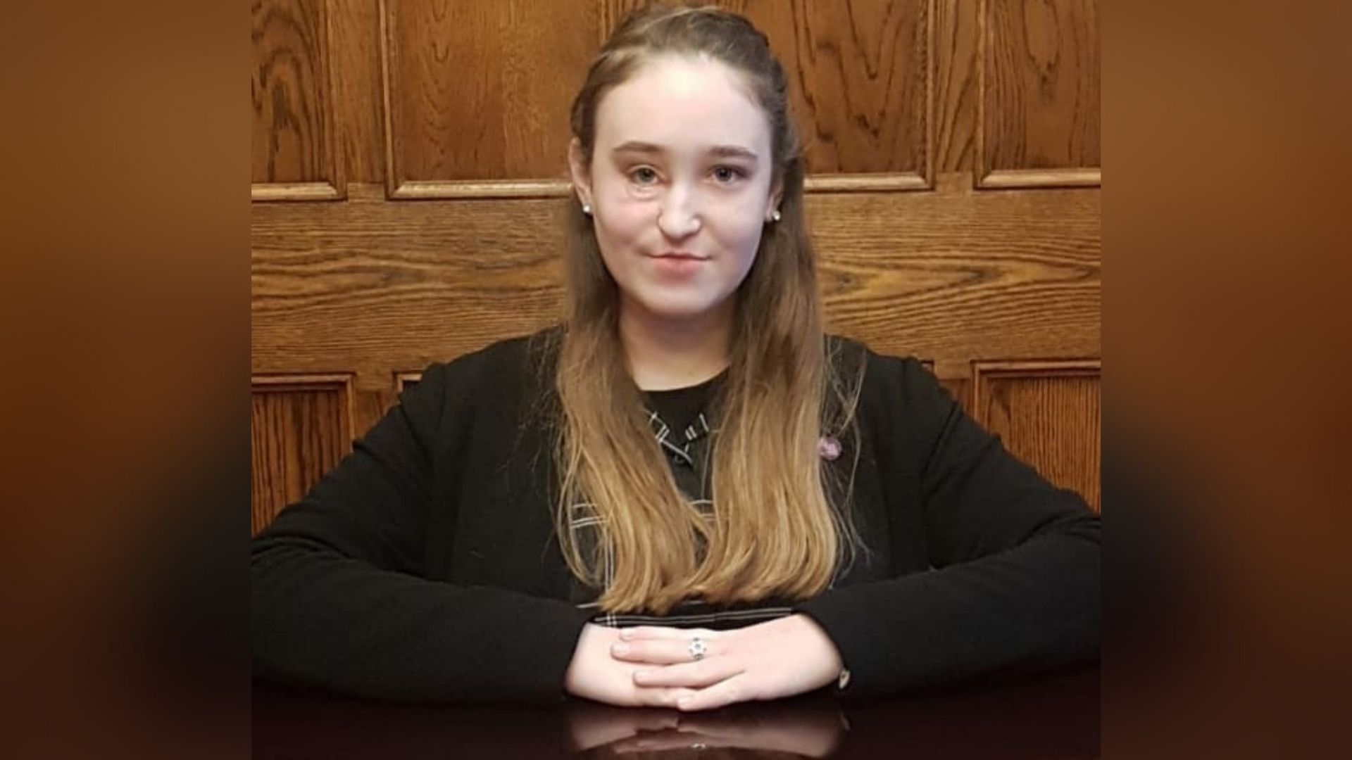 Abigail, a 16-year-old white girl who has long light brown hair and facial scarring caused by the removal of a tumour. She's wearing a black check dress, a black cardigan and sitting at a table with her hands crossed in front of her.