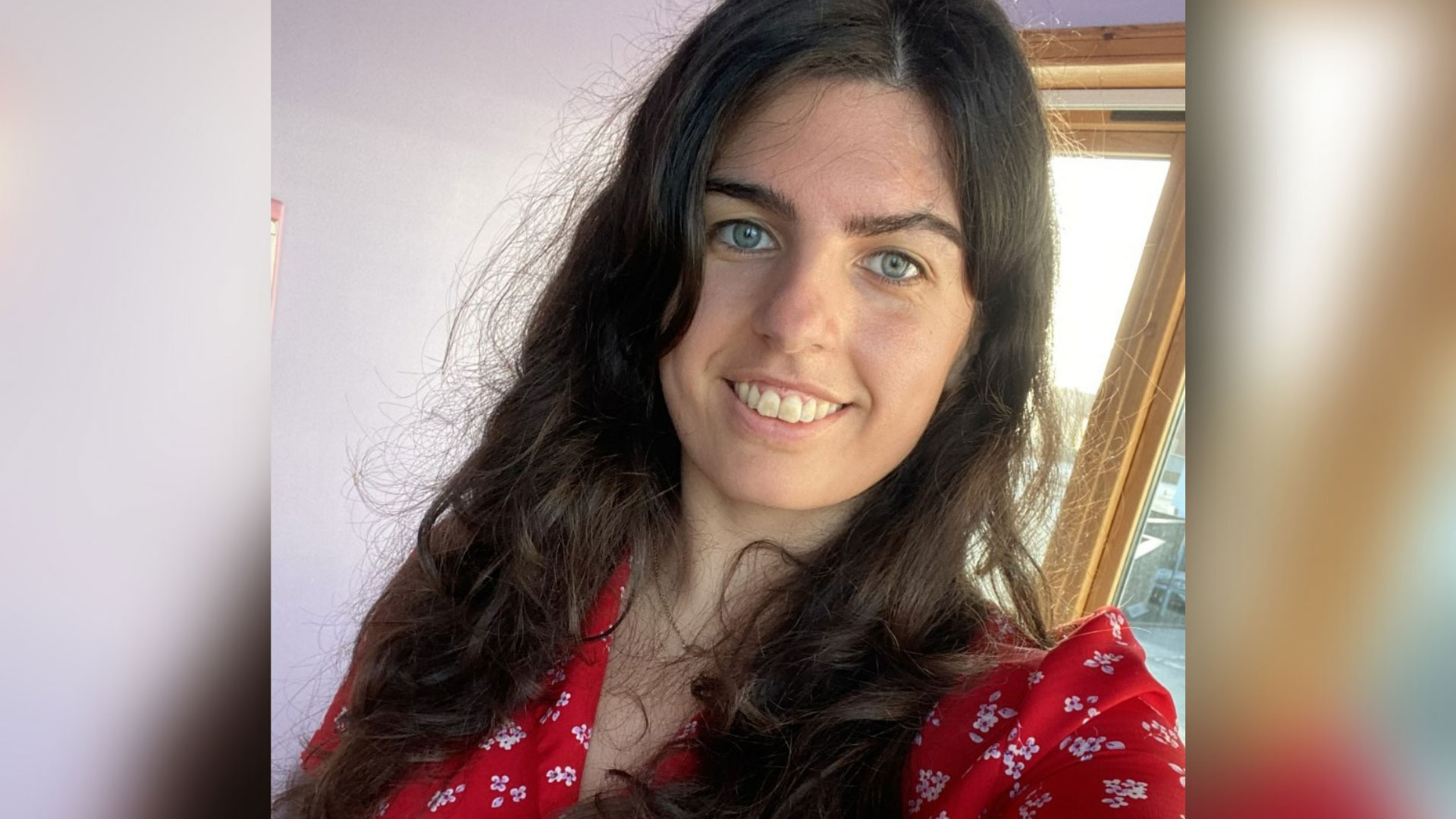 Campaigner Sophie is white with long dark brown hair and a facial visible difference. In this photo she wears a red dress with white flowers on it and the photo captures her head and shoulders.