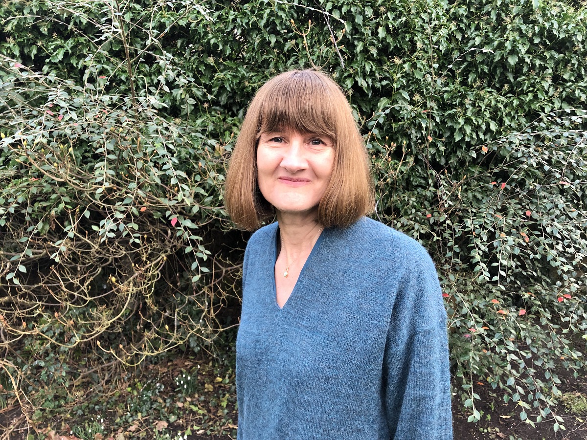 Heather Blake, a woman with shoulder length brown hair and a fringe, standing in front of some bushes