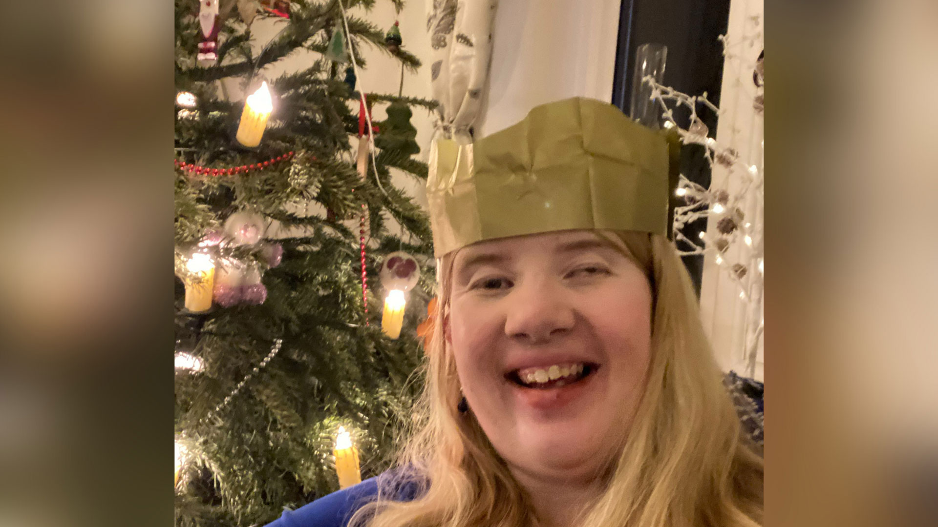 Campaigner Naomi is a white woman with a visible difference and long blonde hair. She is wearing a gold Christmas cracker hat and stood in front of a decorated Christmas tree