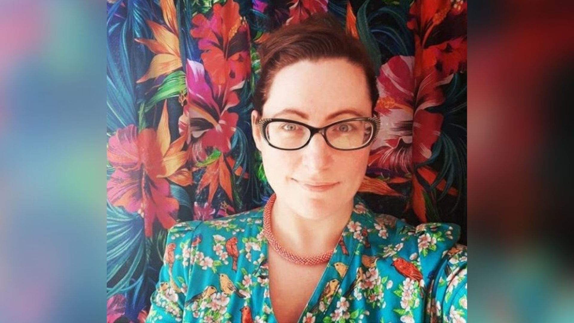Rebecca, a woman who has curly brown hair pulled back from her face. She is wearing a floral print jacket and glasses and looking at the camera.