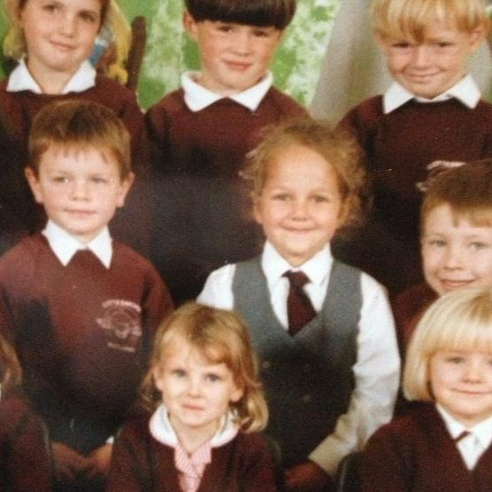 A school photo, with Karen in the middle. She is the only one without a red jumper on, is wearing a white shirt and tie and grey waistcoat. She smiles gently at the camera. Her dark blonde hair is slightly messy in a ponytail.