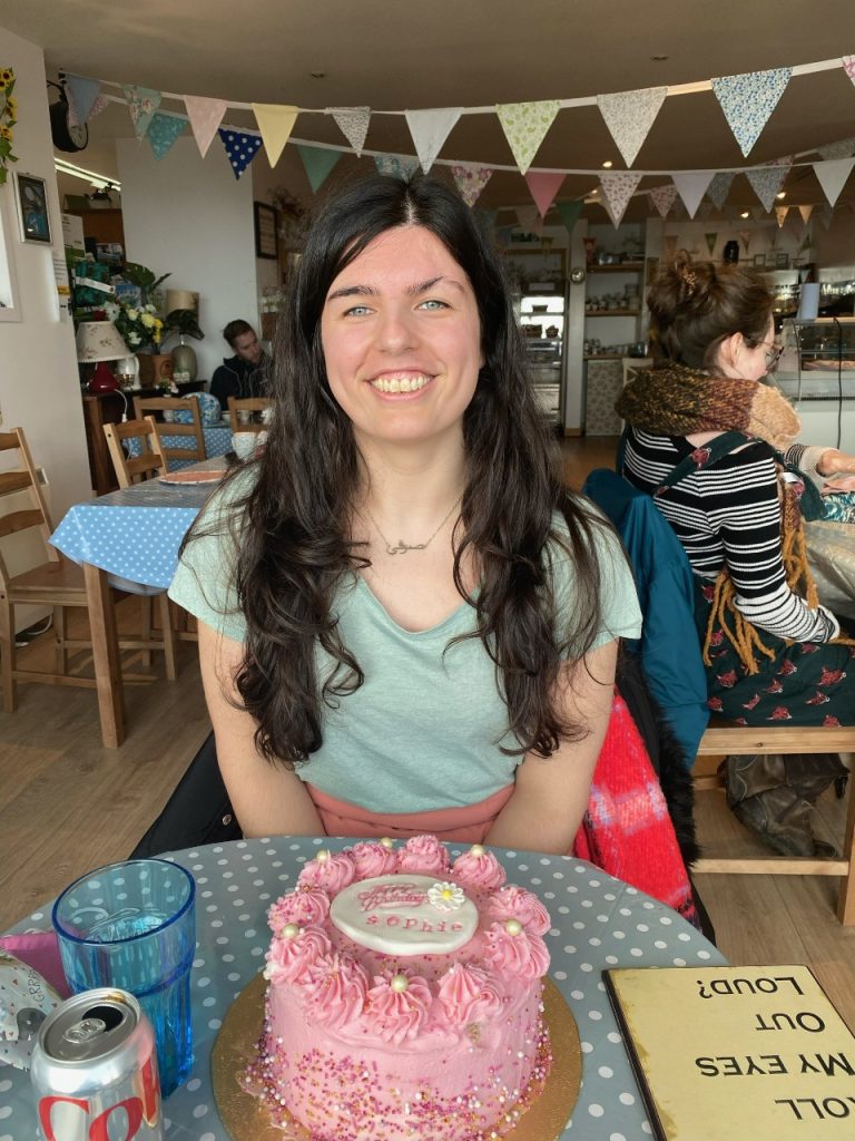 Sophie, sat in a tea room behind a big, pink birthday cake. There is bunting in the background and Sophie is wearing a turquoise top.