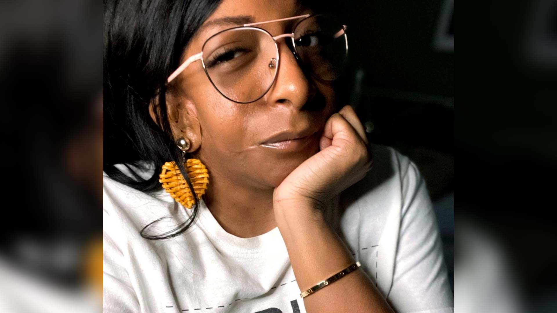 A woman is smiling at the camera with her elbow on a table and her hand propped under her chin. She wears big aviator glasses and big, heart-shaped yellow earrings.