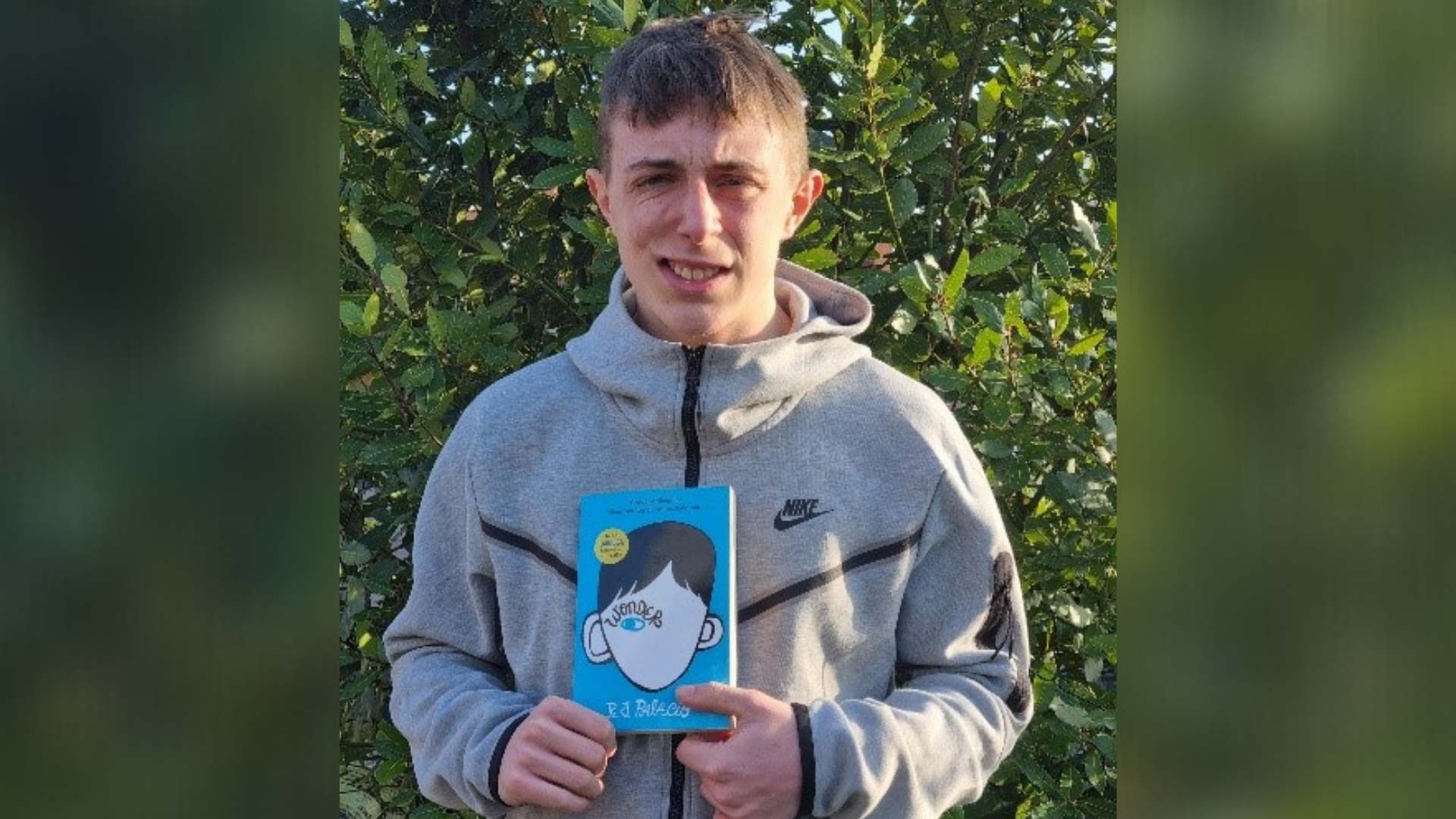 A young man outside, facing the camera and holding a copy of the book "Wonder"