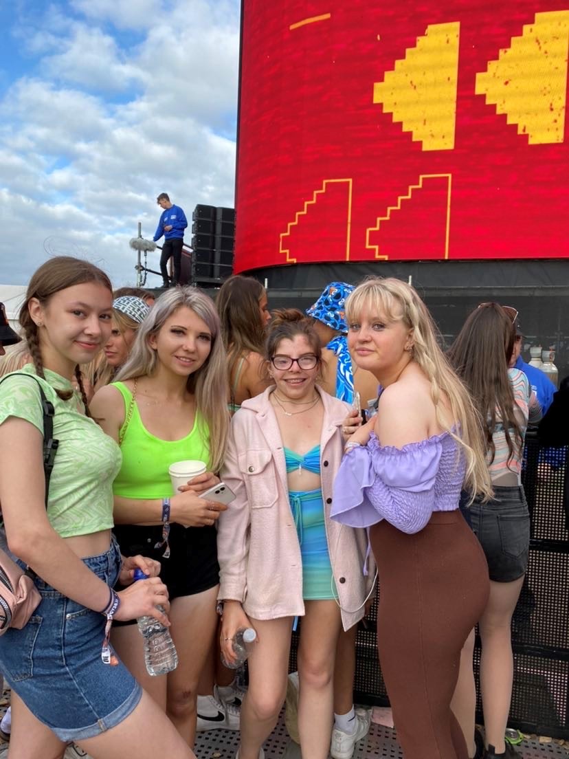 Hannah and her three friends at a music festival. Hannah's in the middle - hair up in a messy bun. She's wearing a very cool two-piece skirt and crop top, in green and blue. She's got a pale pink jean jacket over the top, too.