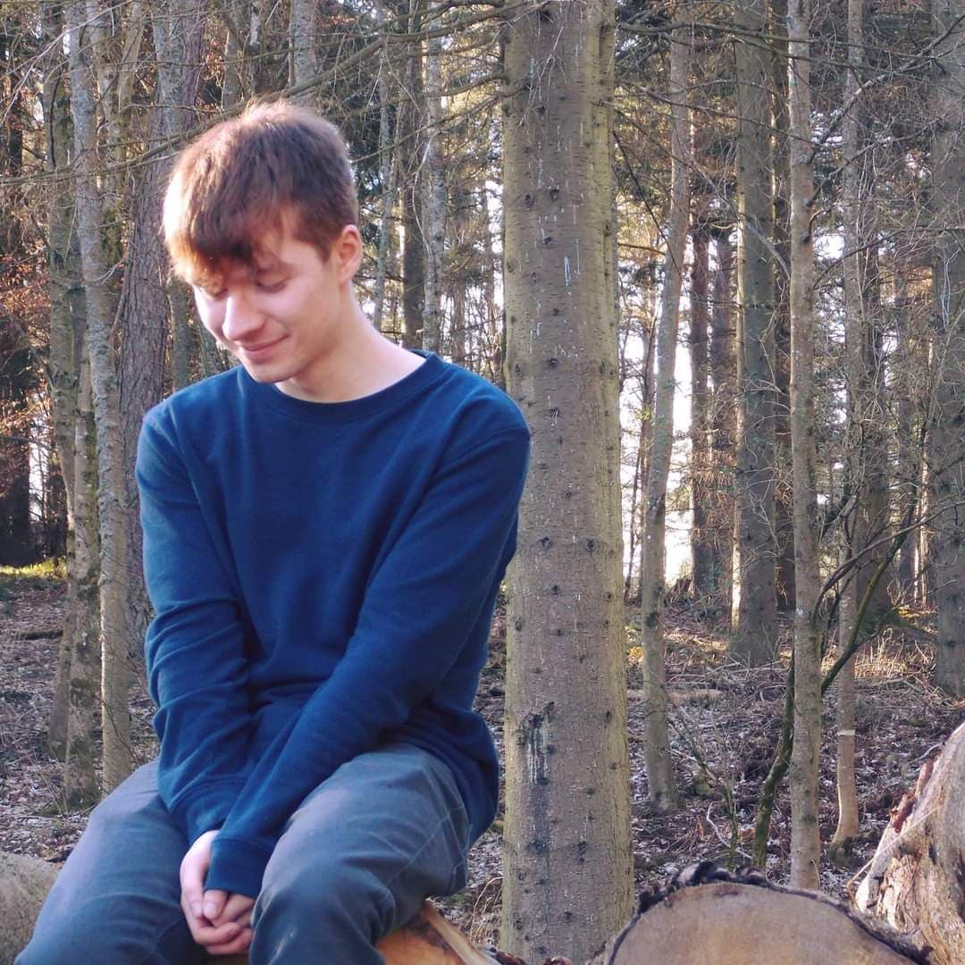 Sam sat in the woods, wearing a blue jumper and jeans. He's sat on some logs and is staring down at the floor, gently smiling to himself.