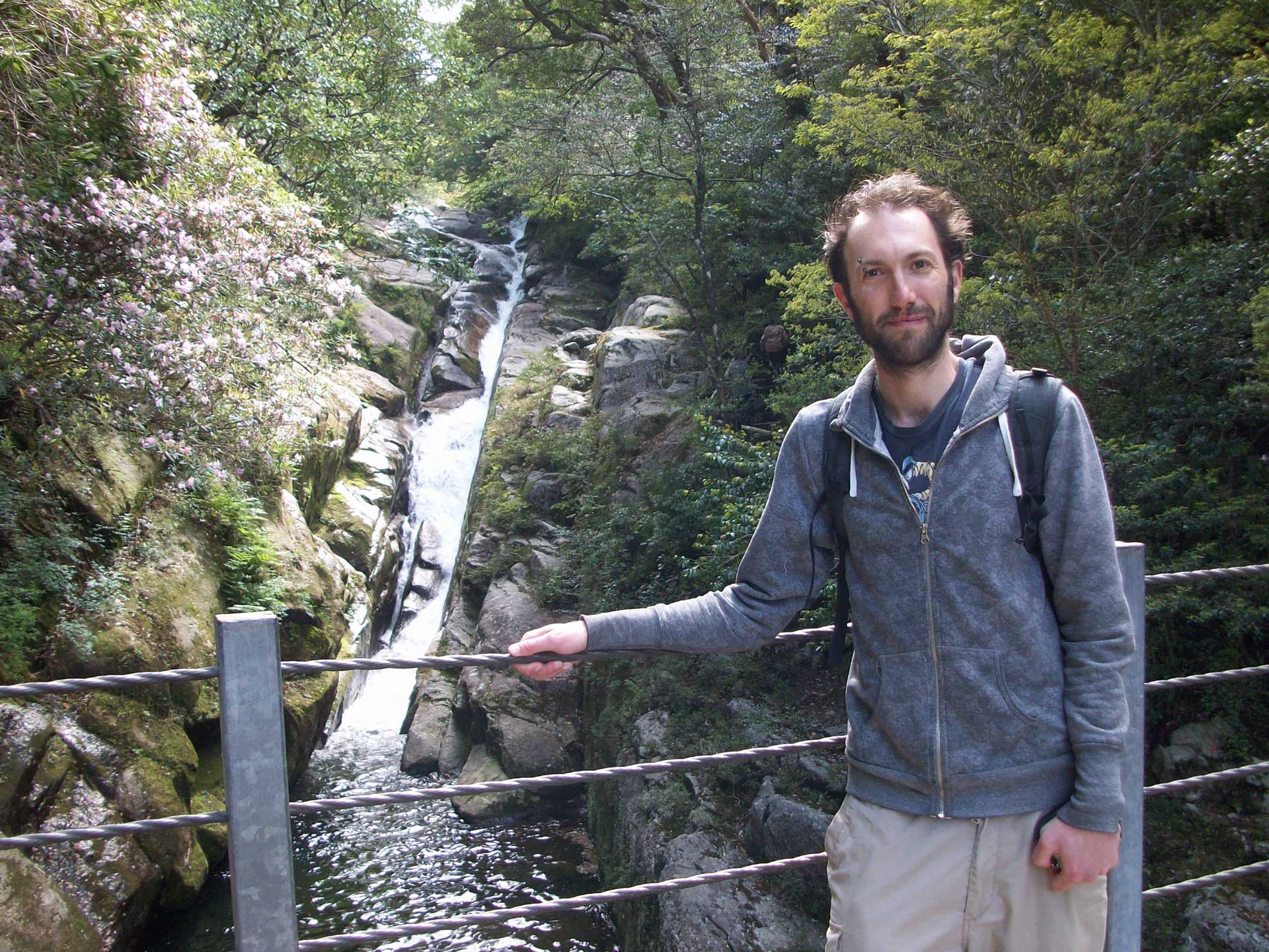 Andrew who is a white male with brown hair and a beard, with a visible difference and an eyebrow piercing, wearing a blue t-shirt, darker blue hoodie and beige trousers, stands next to a waterfall in Japan