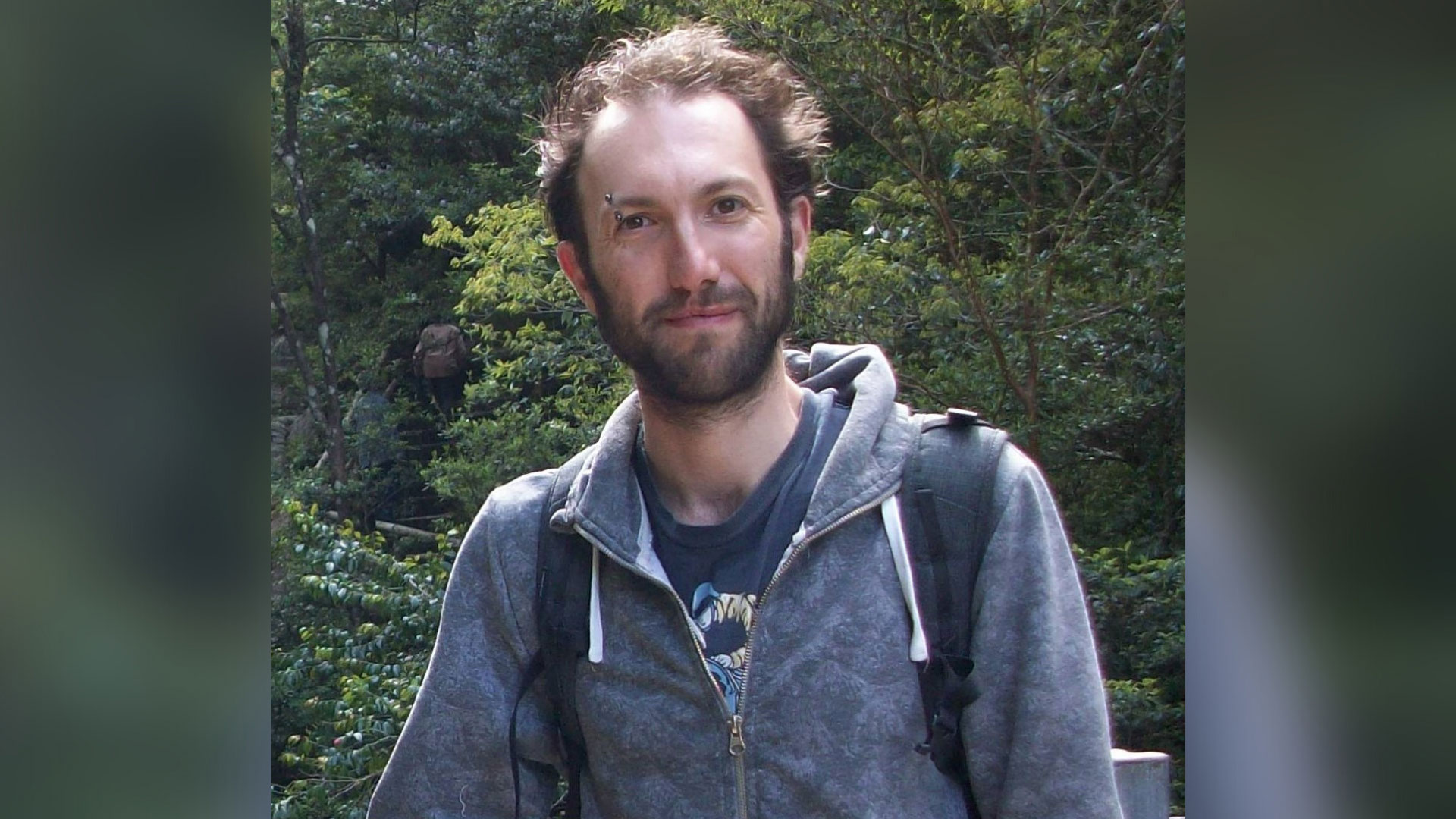Andrew is a white male with brown hair and a beard, with an eyebrow piercing and visible difference. He is wearing a blue t-shirt with a darker blue hoodie over the top.