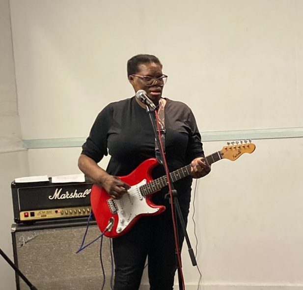 Chrissie is a black woman wit burn scarring on her face and body. She is rocking out with an electric guitar and singing with her band at rehearsal.