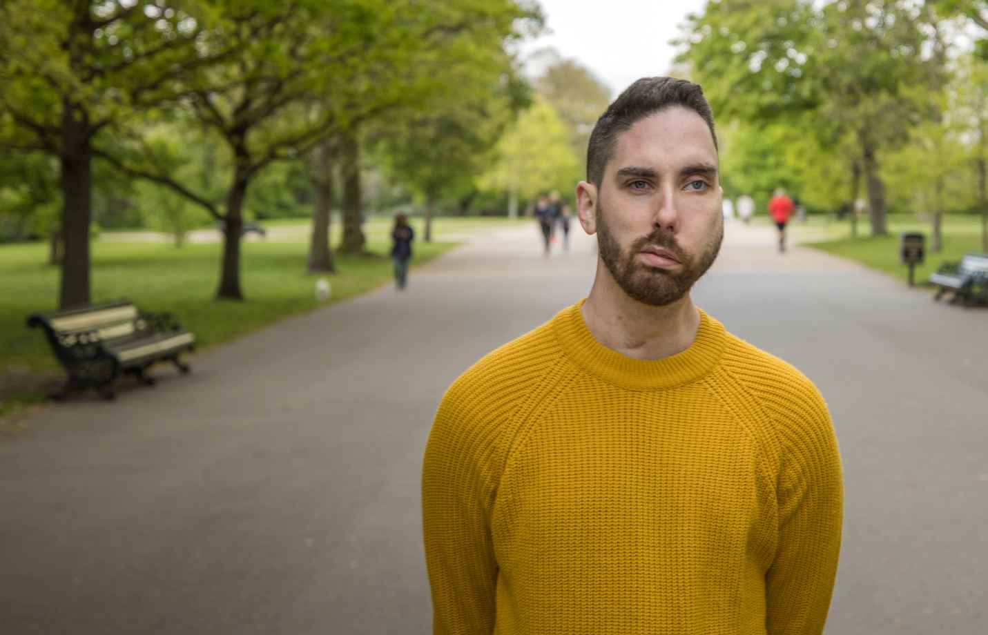 A man with a facial visible difference wearing a yellow jumper, standing expressionless in a park as people walk by.