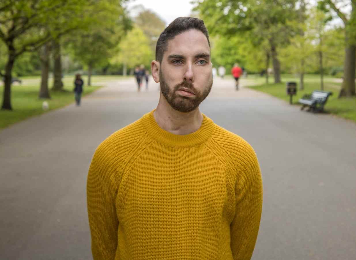 A man with a facial visible difference wearing a yellow jumper, standing expressionless in a park as people walk by.