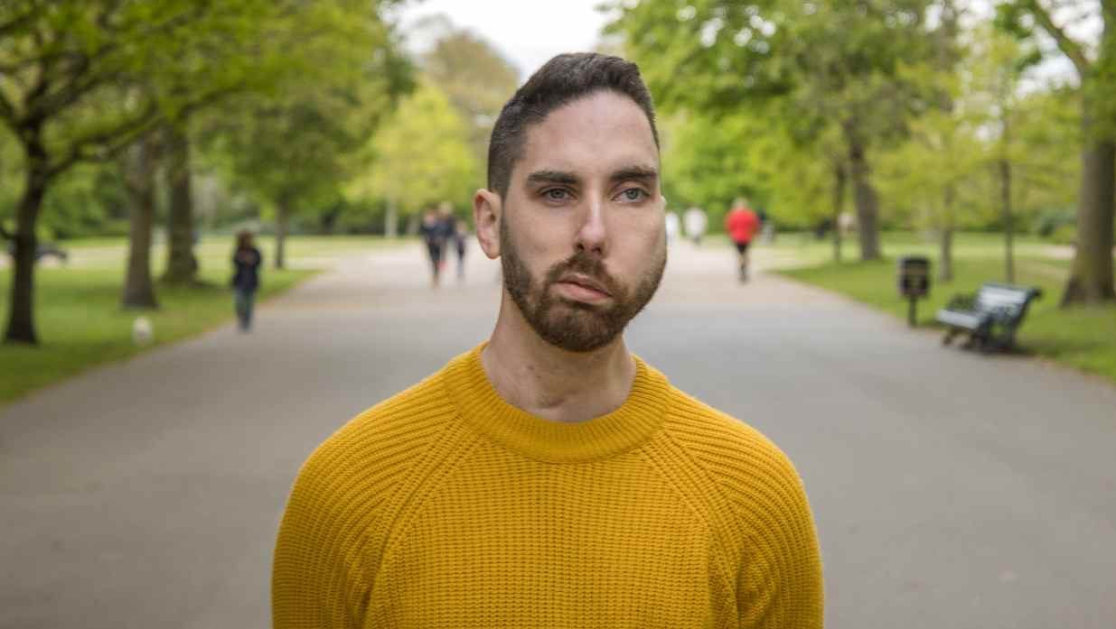 A man with a facial visible difference wearing a yellow jumper, standing expressionless in a park as people walk by.