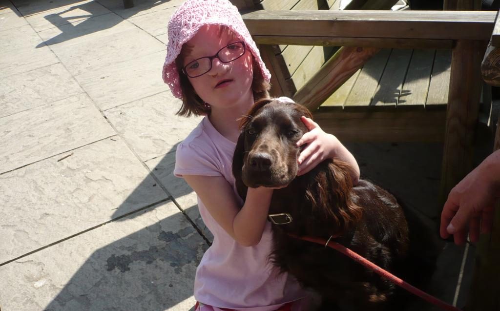 Sophie as a child, hugging her brown cocker spaniel. She wears a pink top, pink crochet hat and pink trousers with apples on them.