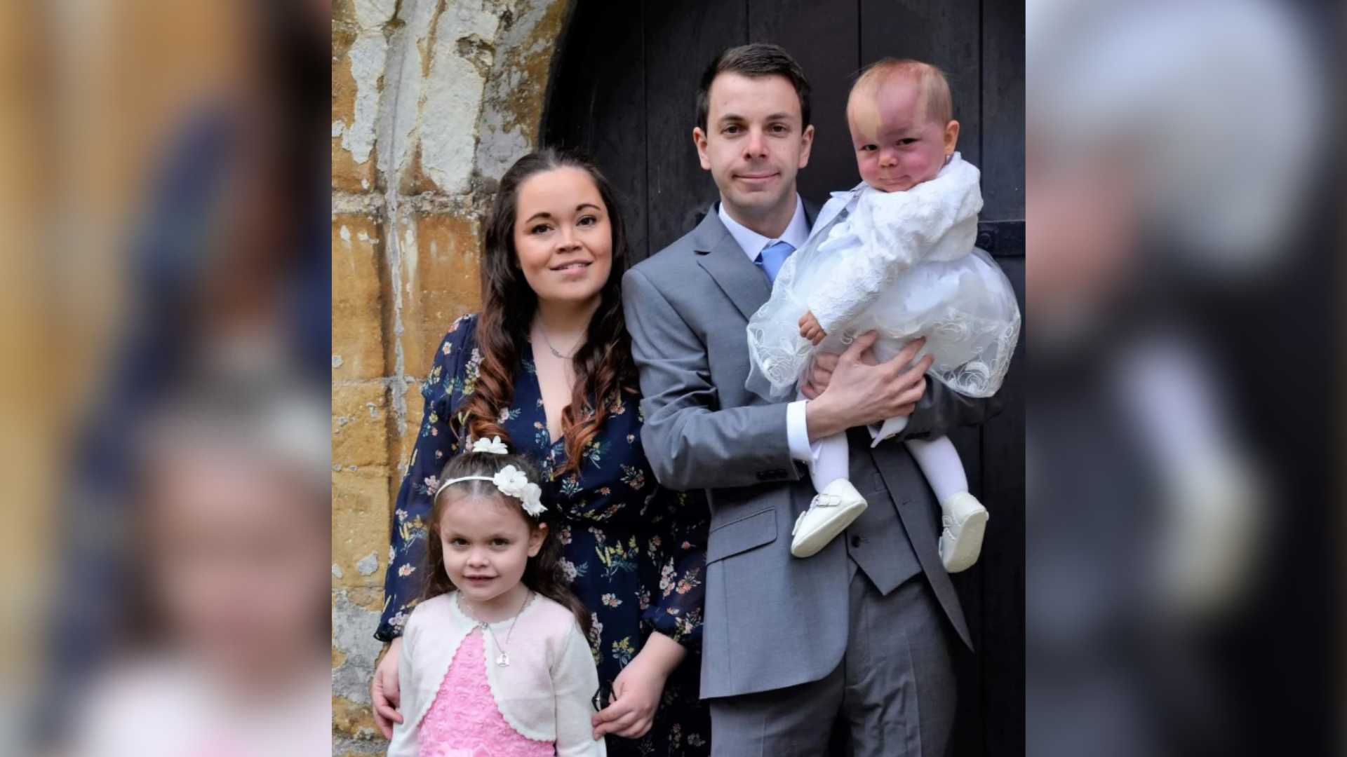 Alice, Dan and their daughters Chloe and Lily at a wedding. They're a brunette family, stood in front of a church door. Lily is held high in her dads arms, smiling cheekily at the camera.
