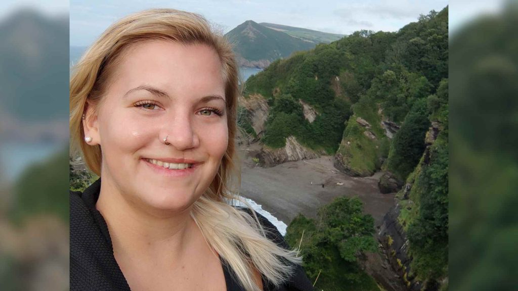 A blonde woman with a cleft scar takes a selfie with a view of a beach and large rocks covered in greenery 