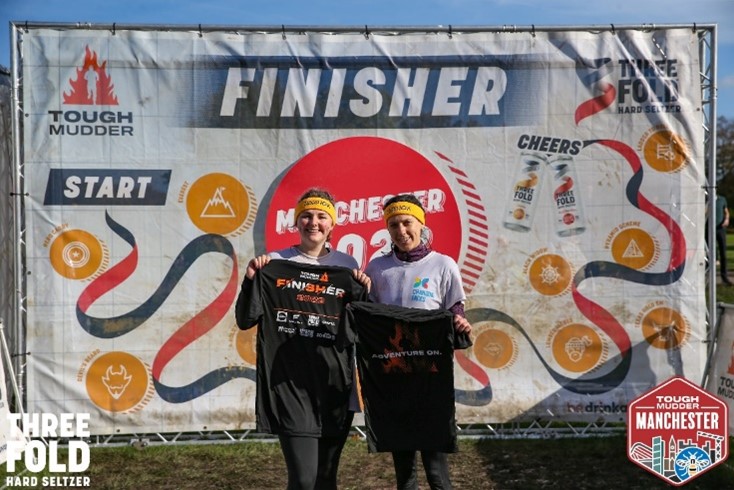 Two women in Changing Faces t-shirts stand in front of a Tough Mudder finish sign