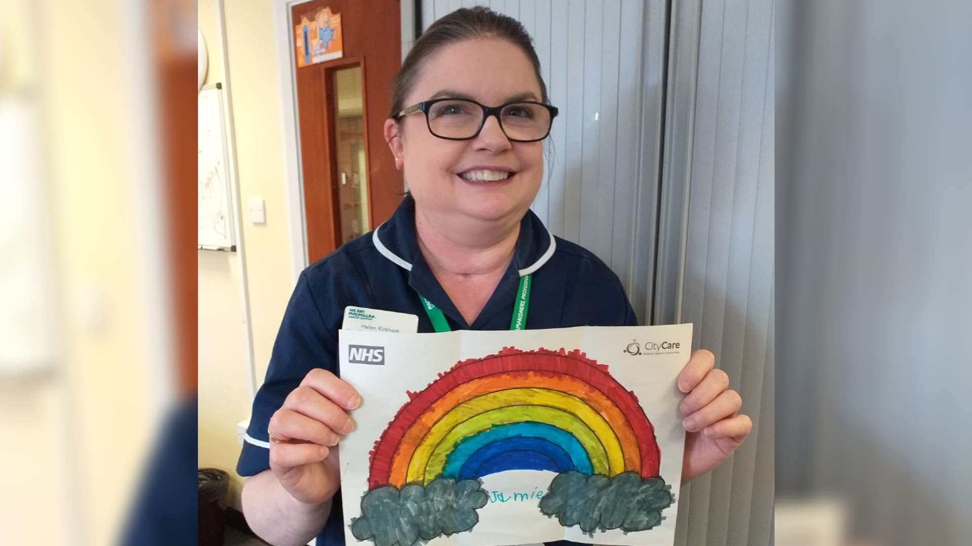 Woman in nurse's uniform with tracheostomy scar stands holding a drawn picture of a rainbow