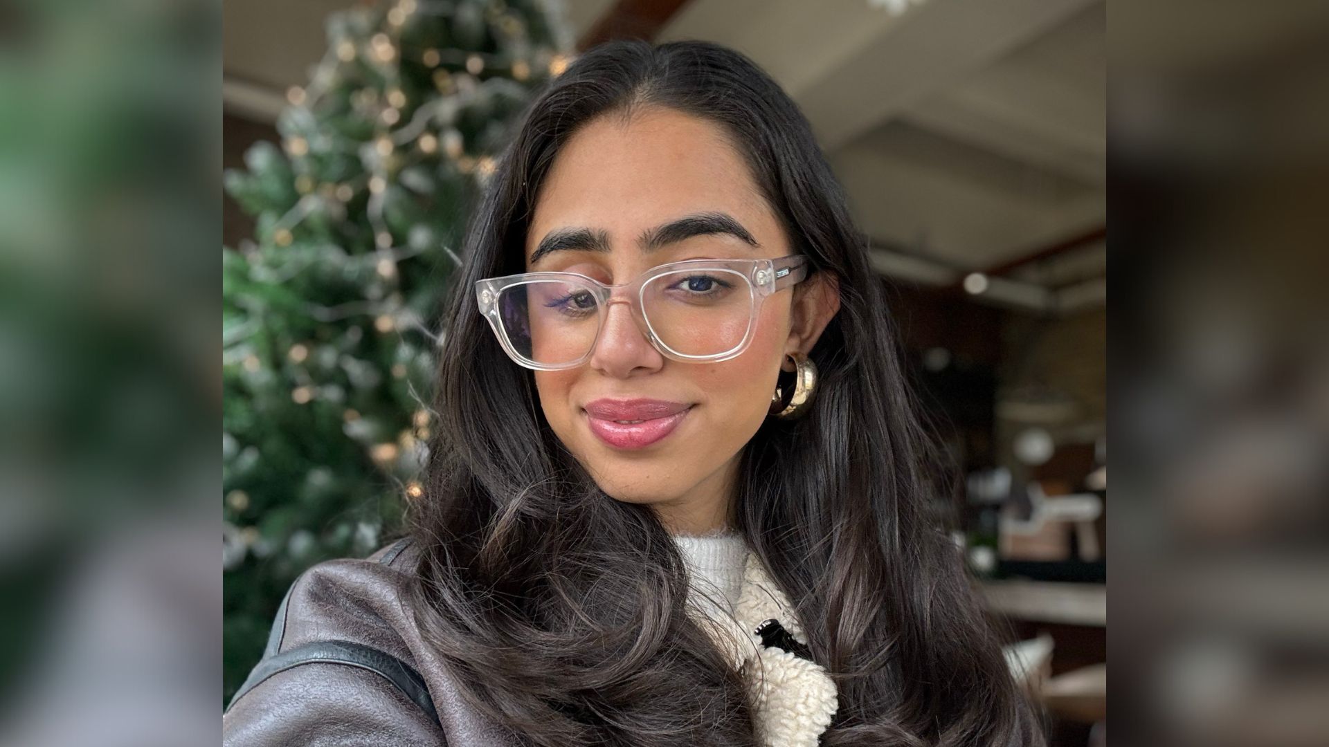 Anahita, a young woman in her 20s, wears glasses and a black jacket and smiles at the camera.