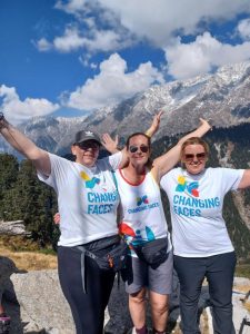 An image of three women wearing changing faces t-shirts standing in front of a mountain range smiling in the sun with arms outstretched