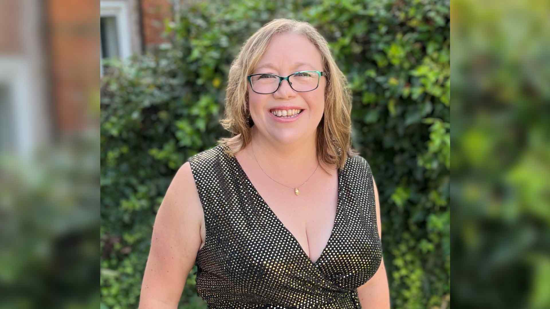 A beautiful photo of Stacey in her garden wearing a black on silver dress. She is wearing green glasses, smiling and has a cleft lip.