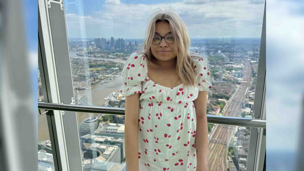 Katie who lives with kyphosis scoliosis, stands by a window smiling at the camera. The view out the window is that of a city from high up.