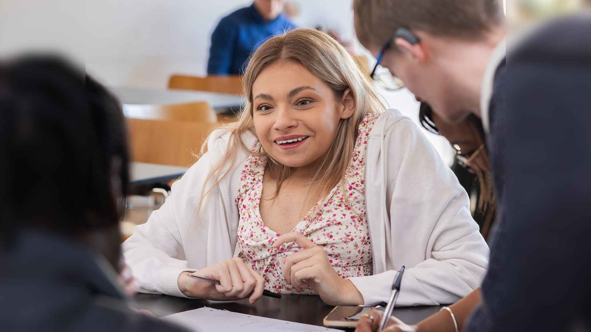 Katie has kyphosis scoliosis. She sits at a table talking to other Changing Faces campaigners. The camera is focused on her.