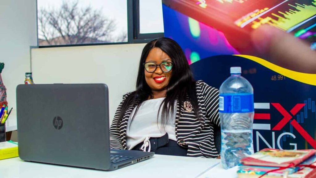 Gillian is a black woman who has Parry-Romberg Syndrome. She sits behind a desk with a laptop in front of her. Gillian smiles at the camera and wears a white t-shirt with a striped cardigan.