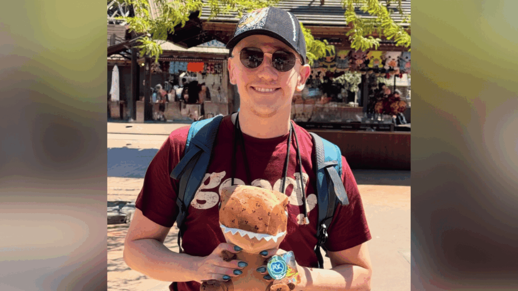 Rob smiles at the camera wearing a baseball cap and sunglasses. He is holding a plush dinosaur toy. Rob is an LGBTQIA+ person with a visible difference.
