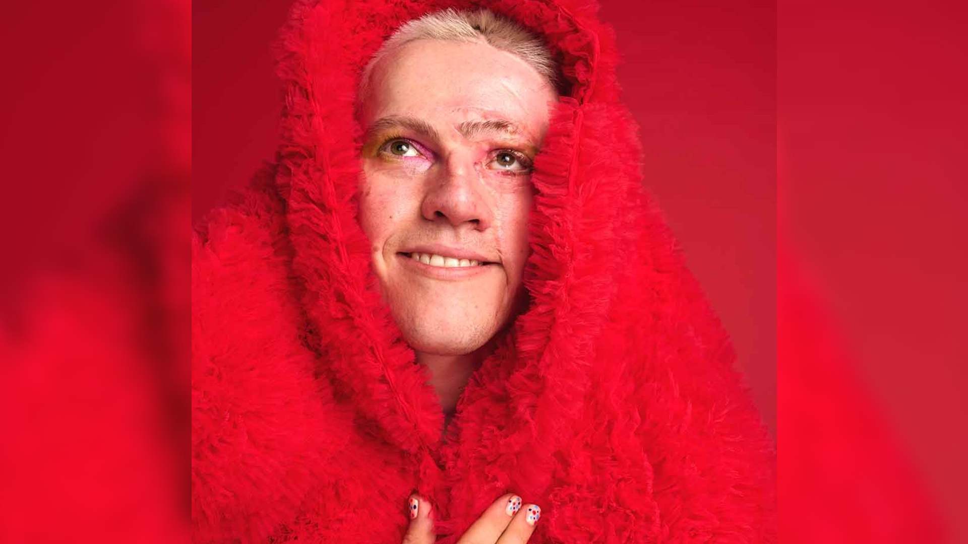 Rob has facial scarring. He is pictured on a red background wearing a red textured coat. His hand is on his chest and his nails are painted with multicoloured dots. This is the hero image for Rob's blog about being a LGBTQIA+ person with a visible difference.