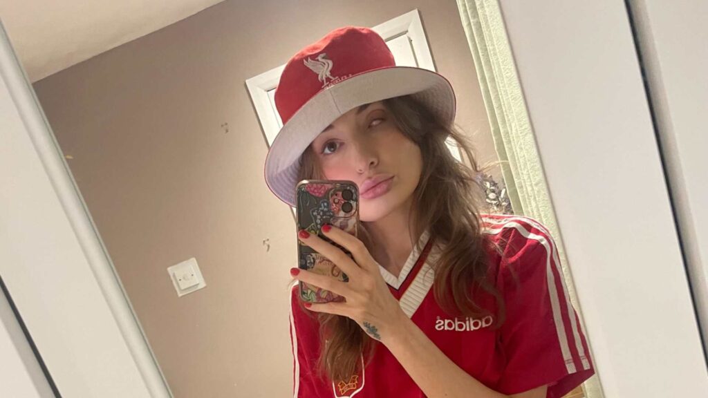 Becky takes a mirror selfie. She's wearing a Liverpool football shirt and hat. Becky is a parent with a visible difference.