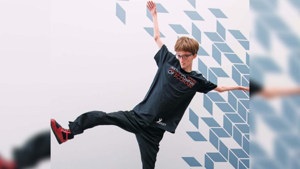 Dylan dances with his arms outstretched and leg lifted to the side. He is wearing a National Youth Dance Company of Scotland t-shirt.