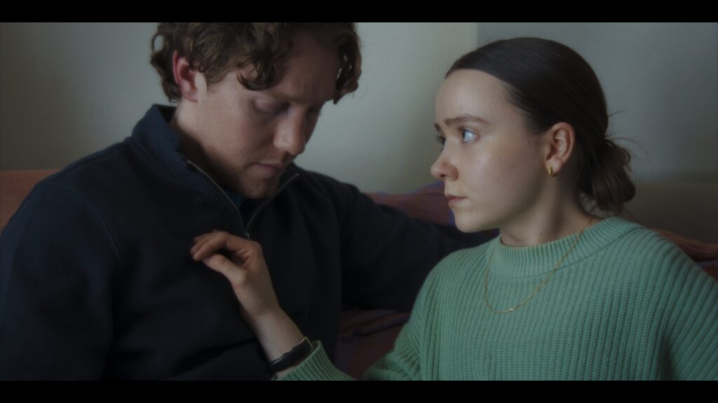 A still from Hugo's film Scratch. A woman and a man are next to each other shown from the shoulders up. The woman looks at the man's face with her hand on the man's chest. The man looks down at her hand. Scratch has helped Hugo with overcoming the shame around his scar.
