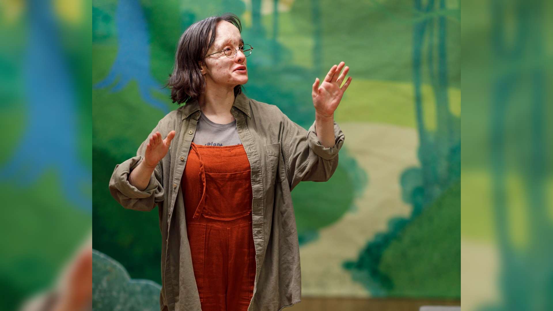 Beau acting. They are looking off to the side, using their hands to emphasise their words. The background is a painted pathway in a forest. Beau is showing that actors with visible differences can take to the stage.
