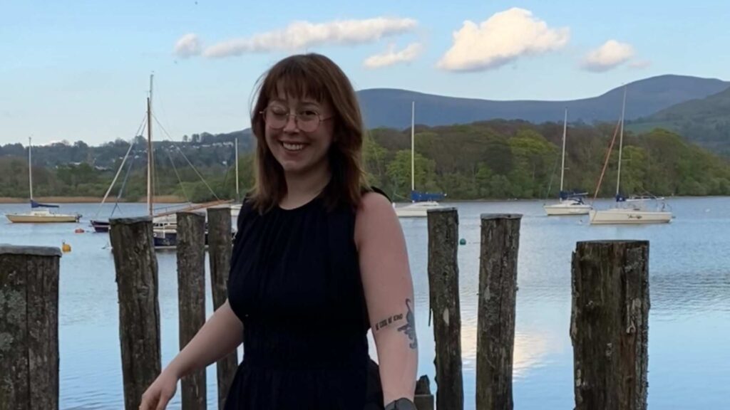 Savannah lives with NF2. She stands on a wooden pier on a lake with boats in the background. One of her eyes is closed due to her condition.