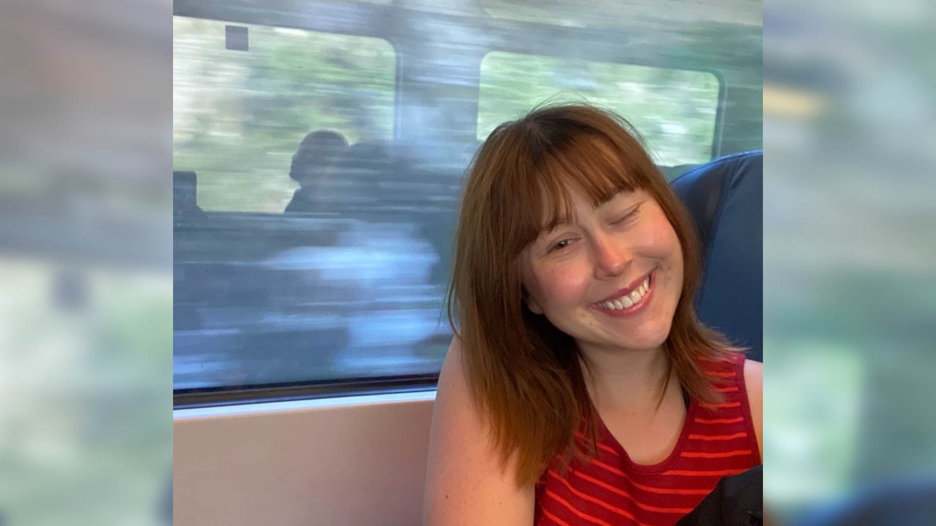 Savannah lives life with NF2. One of her eyes is permanently closed. She smiles at the camera while sat on a train. The view out of the window is blurred from the motion of the train.