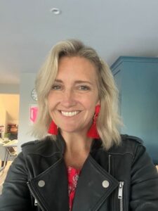 Caroline smiles at the camera wearing a black jacket and red ear rings