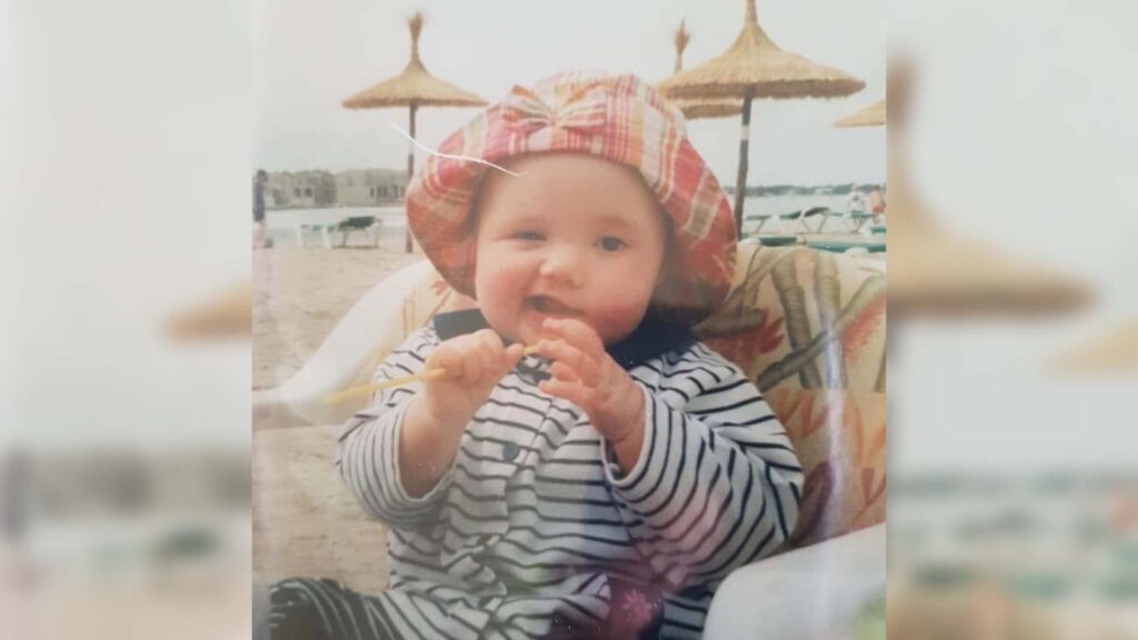Chloe as a baby. She is sat on a beach with straw beach umbrellas in the background. 