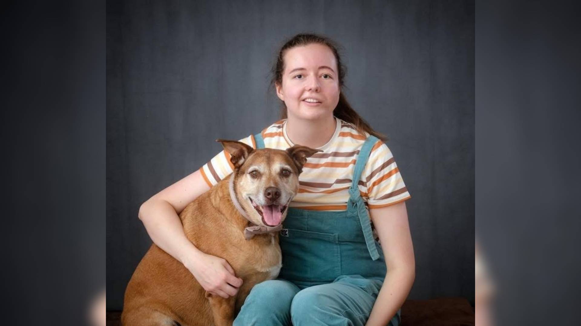 Chloe sits in front of a grey photography background with a brown dog sat next to her. Chloe's arm is around the dog. Chloe has facial palsy.