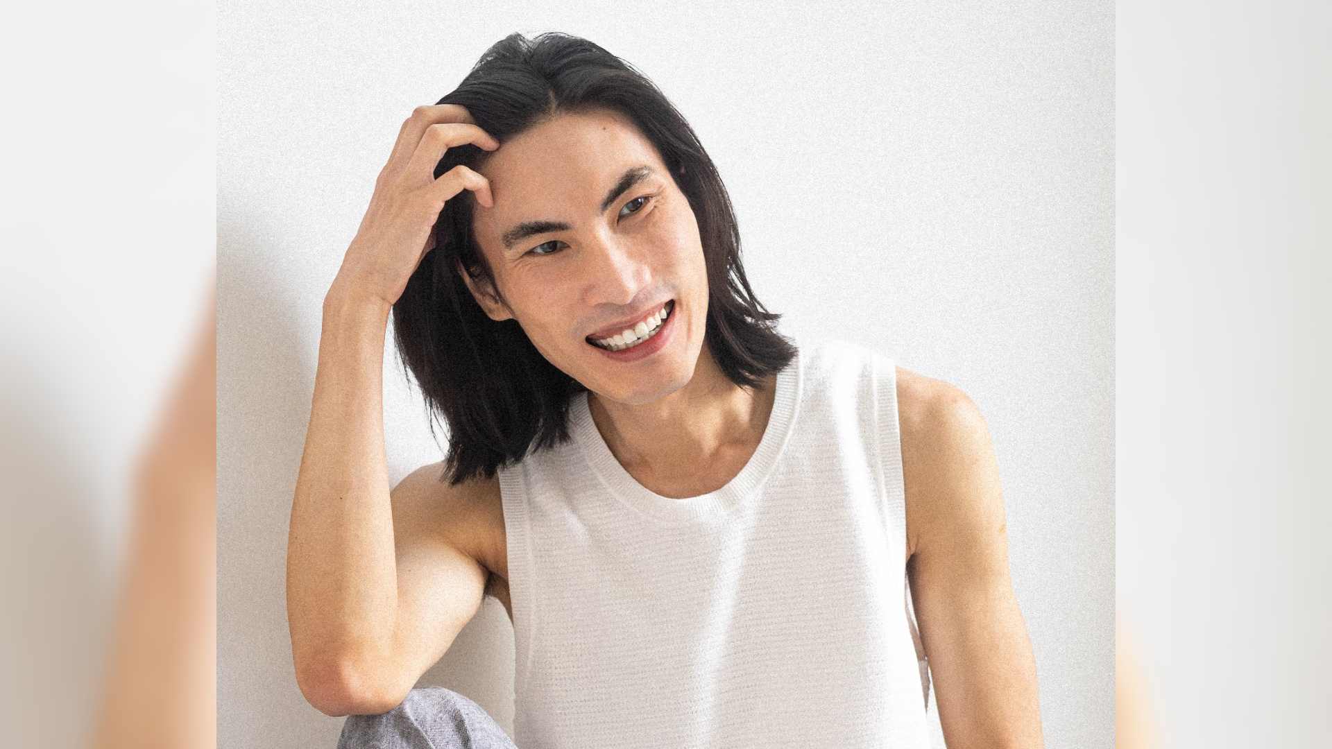Eli is an Asian man with shoulder length black hair. He leans his elbow on his knee with his head leaning against his hand. He smiles looking away from the camera.