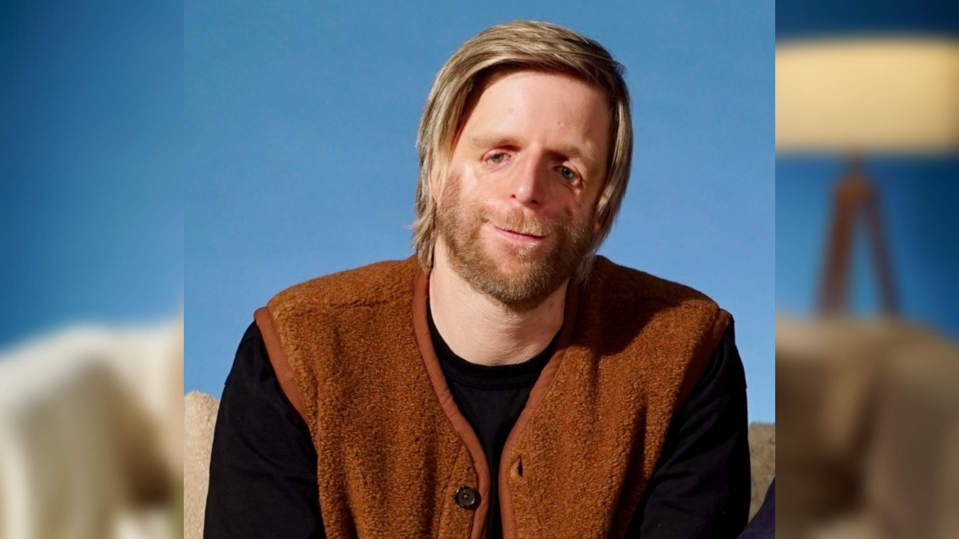 Jono Lancaster is a white man with blonde hair. He has Treacher-Collins Syndrome. He sits on a sofa wearing a black top and brown sleeveless cardigan.