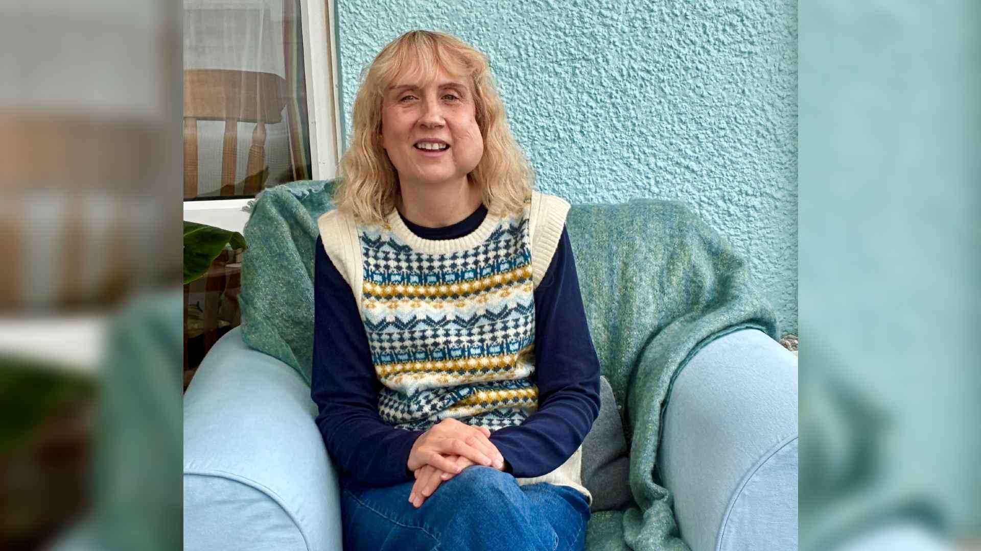 Mary is a blonde-haired woman who has a haemangioma on the left side of her face. She sits in an armchair and smiles at the camera, her hands on her lap.