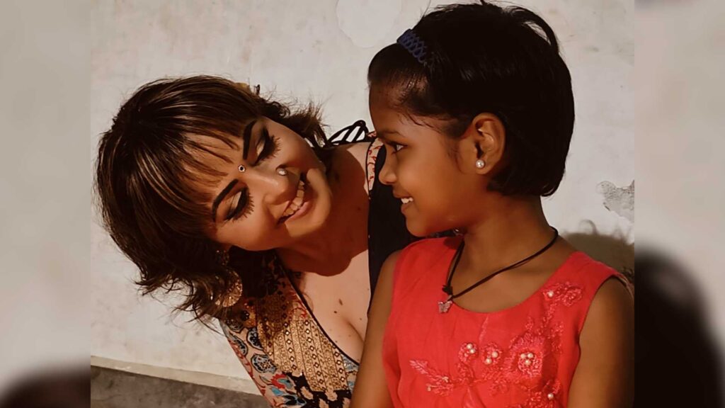 A photo of Ash and a young girl. Ash peaks around the side of the girl with a smile on her face.