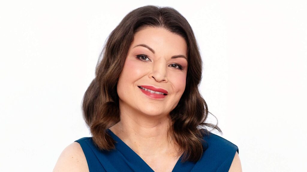 A professional headshot of Vikki. She wears a dark blue dress and smiles at the camera. Vikki has a facial difference after cancer affecting her nose.