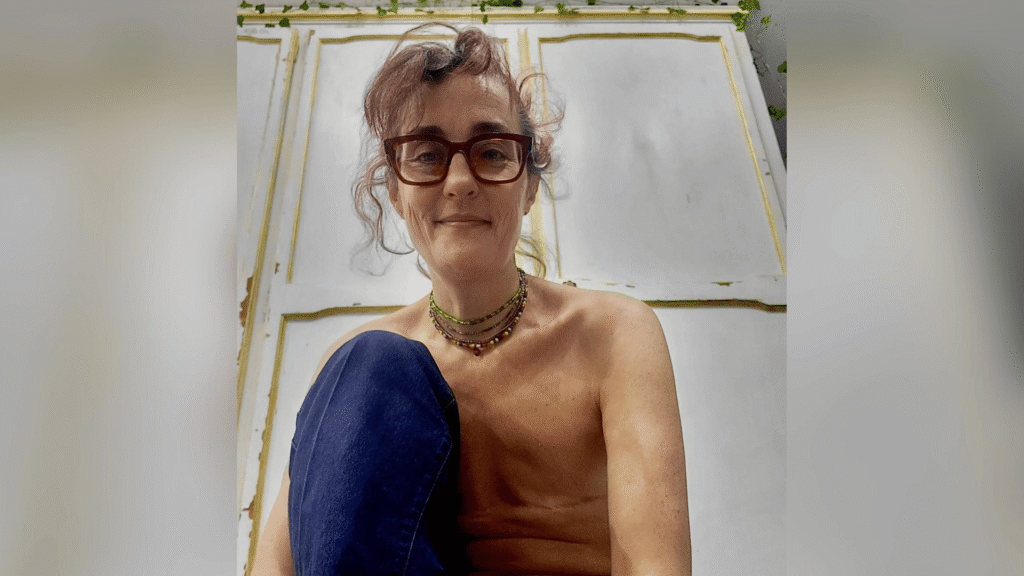 Caroline sits with one leg bent up. Her mastectomy scars and visible.